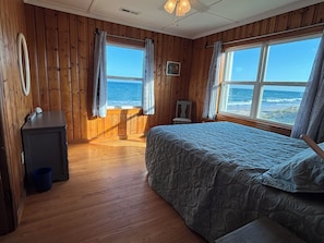 Ein Zimmer aus Holz mit einem Bett, einem Sessel und einem Schreibtisch. Zwei Fenster mit Vorhängen, eines davon bietet Meerblick.