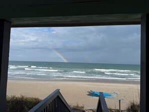 Une vue sur la plage, aperçue à travers une fenêtre ornée d’une balustrade en bois, avec un parasol bleu et un arc-en-ciel dans le ciel.