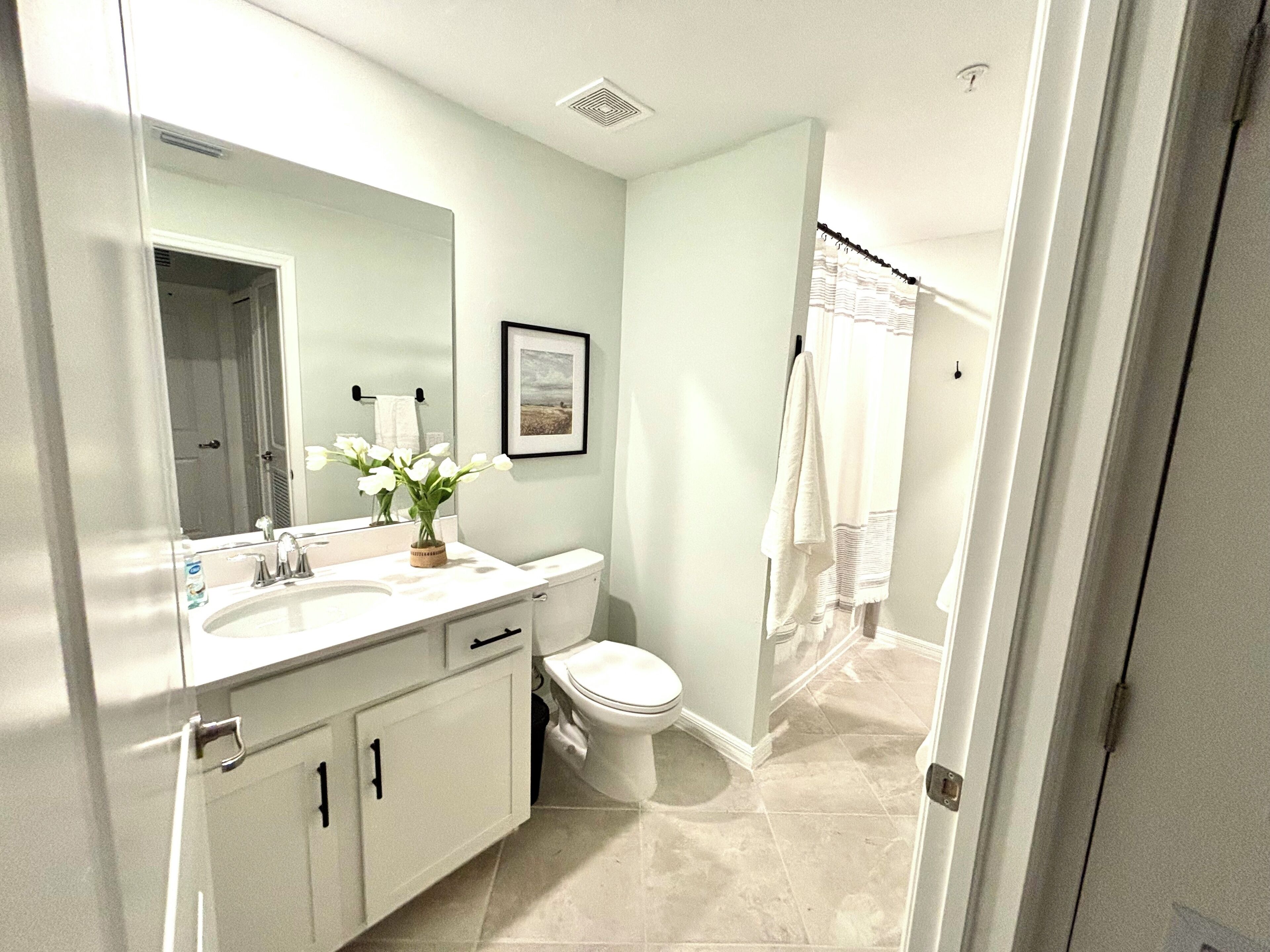 A bathroom with a white sink, mirror, and toilet. There is a framed picture on the wall and a towel rack.