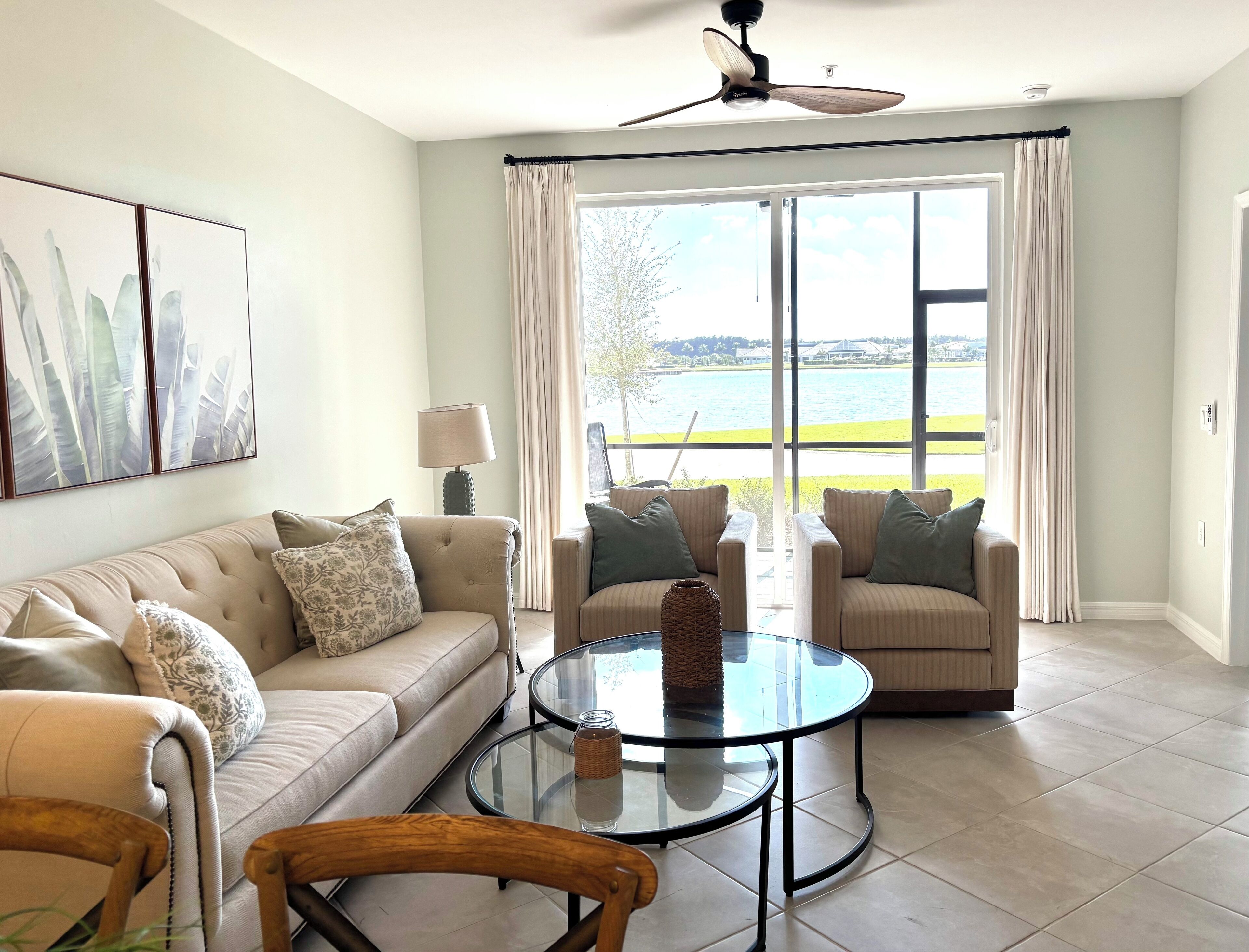 Un salon avec un canapé beige, deux fauteuils, une table basse en verre et un ventilateur de plafond.