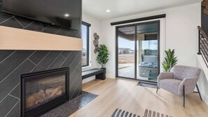 A modern living room with a fireplace, a bench, a chair, and a large window offering a view of the outdoors. - Classy Corner Retreat | Hot Tub + Fireplace (Fort Collins)