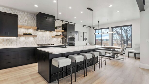 A modern kitchen with dark cabinetry, a central island with bar stools, and a large window providing a view of the outdoors. - Classy Corner Retreat | Hot Tub + Fireplace (Fort Collins)