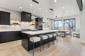 A modern kitchen with dark cabinetry, a central island with bar stools, and a large window providing a view of the outdoors. - Classy Corner Retreat | Hot Tub + Fireplace (Fort Collins)