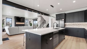 A modern kitchen with a central island, dark cabinetry, and a staircase leading to an upper level. - Classy Corner Retreat | Hot Tub + Fireplace (Fort Collins)