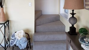Uma escadaria que leva a um segundo andar, um quadro emoldurado na parede, um abajur sobre uma mesa e um vaso com folhagens.