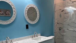 A bathroom with a blue wall, two oval mirrors, a white sink with three faucets, a toilet, and a shower with a glass door.