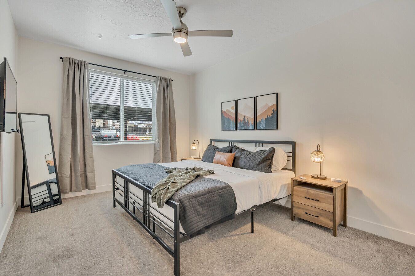 A bedroom with a bed, bedside table, nightstand, ceiling fan, window with curtains, and artwork on the wall.