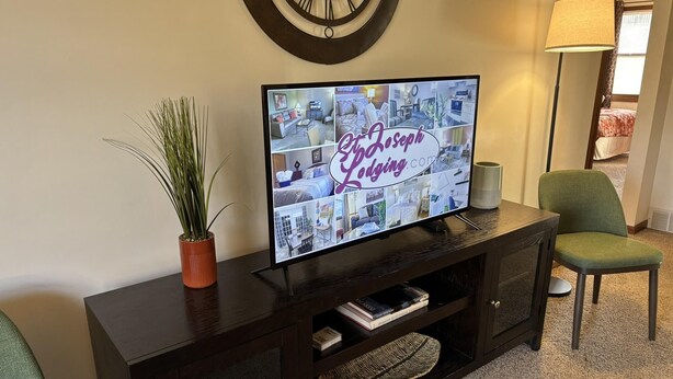 Een televisie waarop de website van Joseph's Lodging wordt weergegeven, een donkere houten entertainmentkast met een plant, een lamp en een groene stoel.