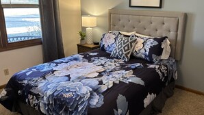 Een slaapkamer met een groot bed, een nachtkastje met een lamp, een lijst met een afbeelding aan de muur en een raam met gordijnen.