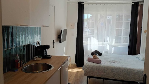 Ein kompaktes Hotelzimmer mit Küchenzeile, einem Bett mit gemustertem Überwurf, einem Fenster mit Vorhängen und Blick auf umliegende Gebäude.