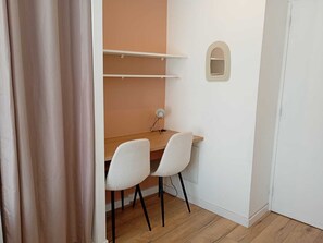 Une petite pièce avec un bureau en bois, deux chaises blanches, un petit miroir rond et une porte blanche.