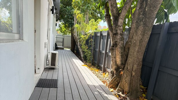 Una terraza de madera que lleva a una casa de fachada blanca, un respiradero metálico y una valla de madera.