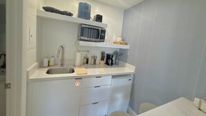 Cocina compacta con lavamanos, microondas y cafetera. Hay armarios debajo de la encimera y un refrigerador pequeño.