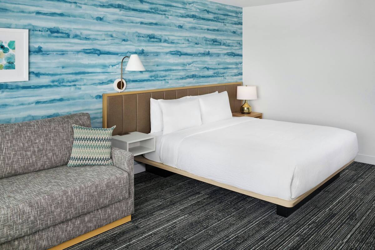 A hotel room with a bed, a sofa, a nightstand, a lamp, and a wall with a blue abstract design.