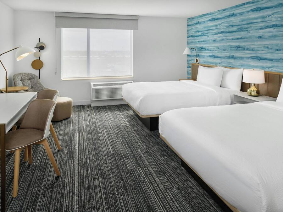 A hotel room with two beds, a desk with a chair, a small table, and a large window.