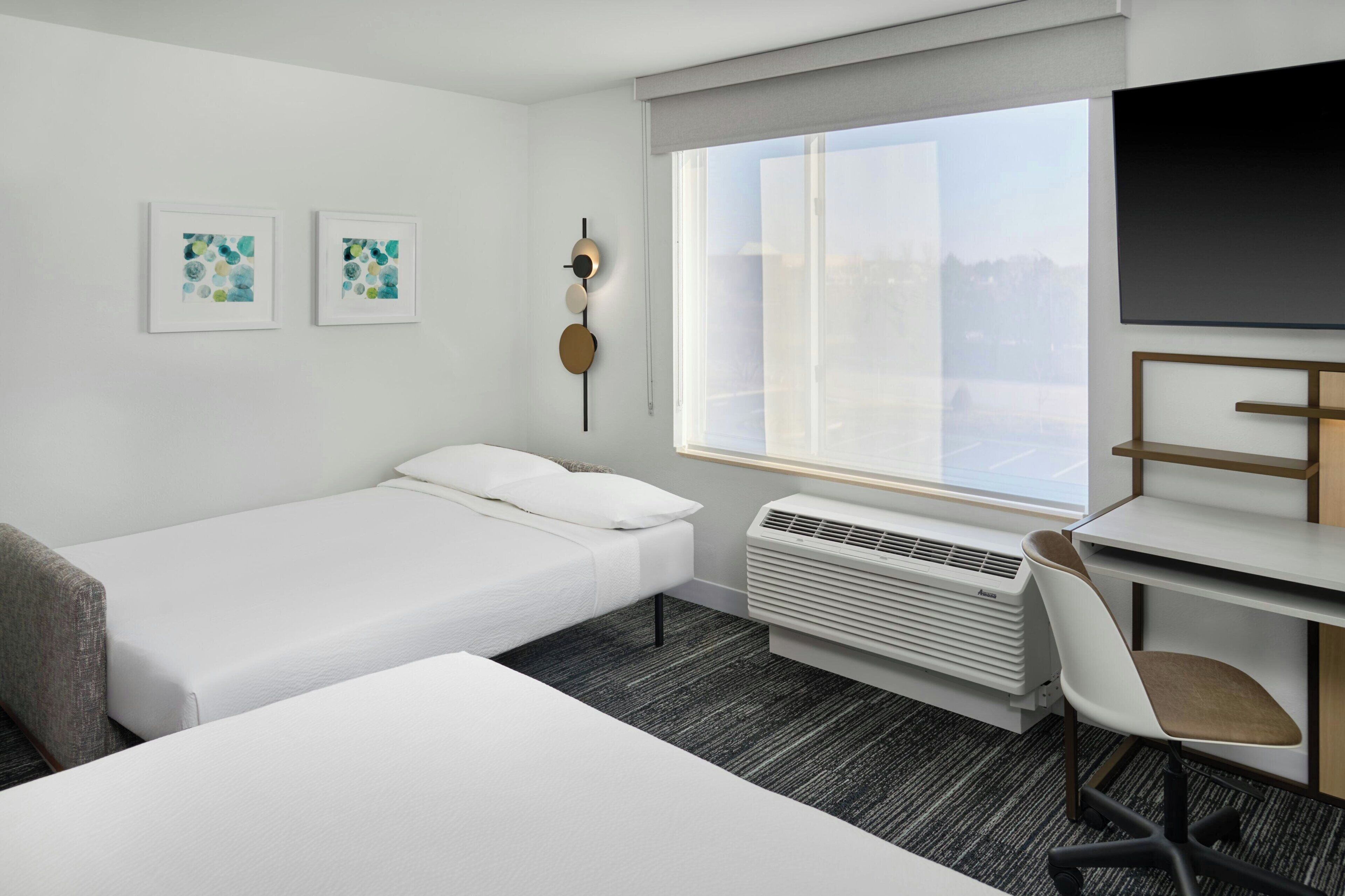 A hotel room with a bed, a desk with a chair, a TV, a window with a view, and two wall-mounted artworks.