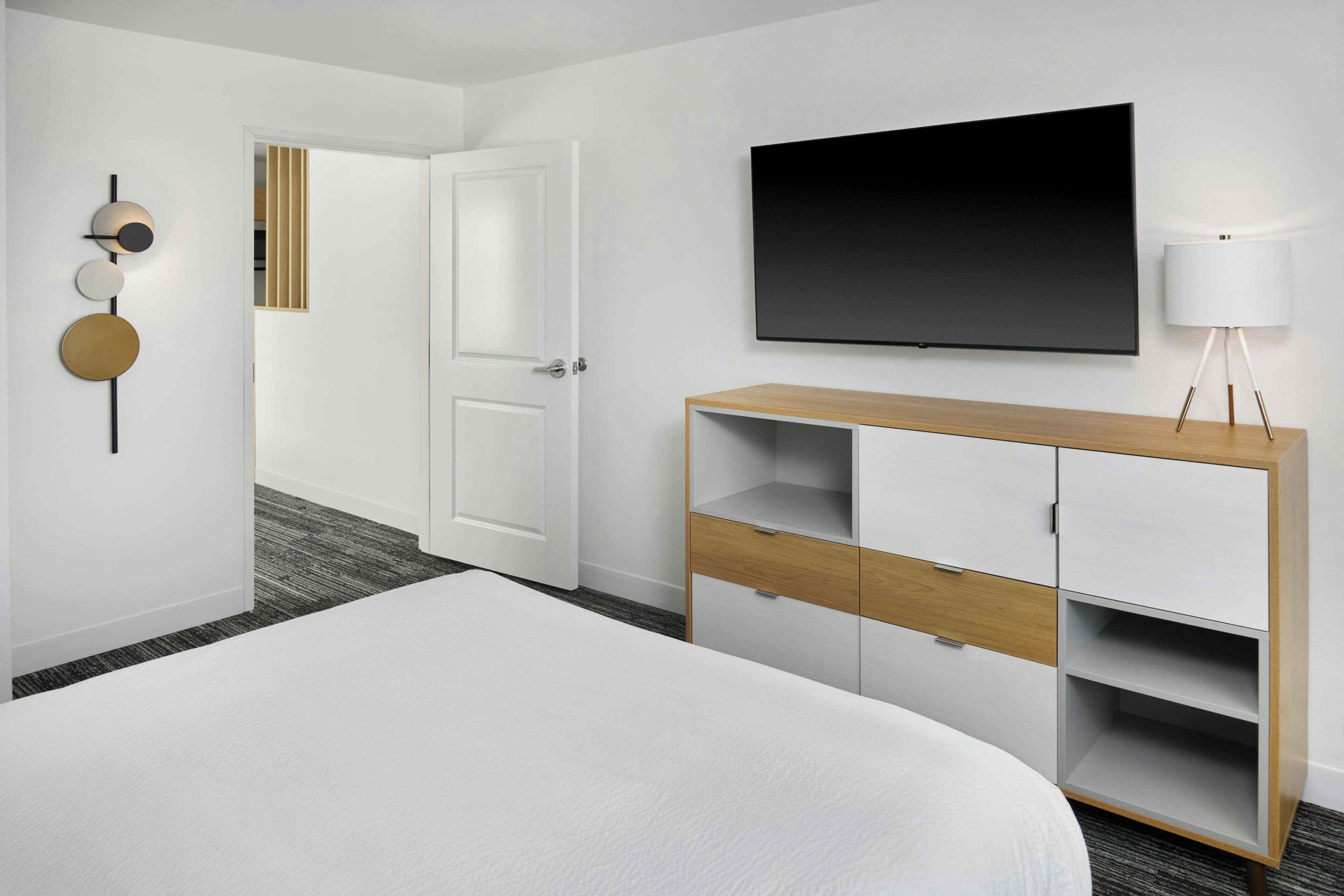 Ein Hotelzimmer mit einem Bett, einem an der Wand befestigten Fernseher, einem Holzschrank und einer Tür zu einem weiteren Raum.