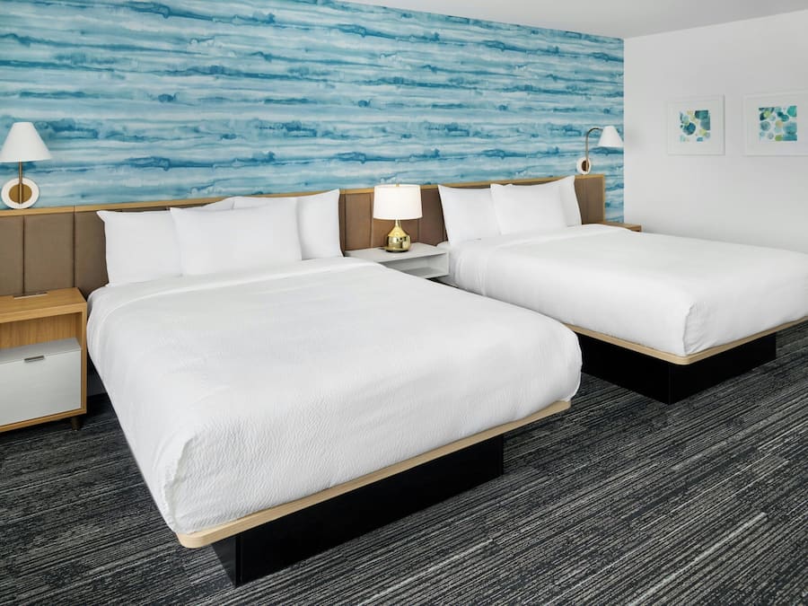 Habitación de hotel con dos camas, una mesita de noche con lámpara, un cajón y una pared azul con textura.