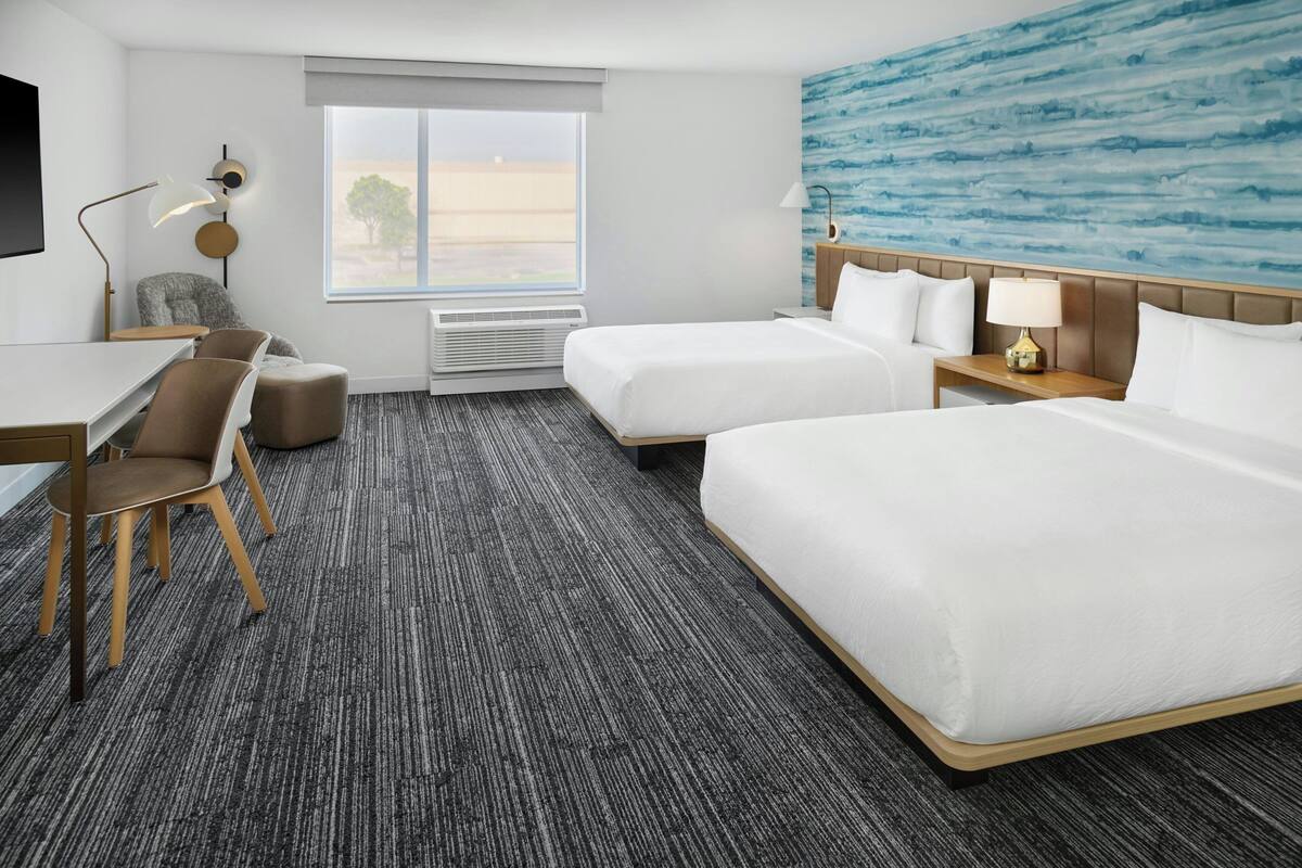 A hotel room with two beds, a desk with chairs, a TV, and a large window with a view.