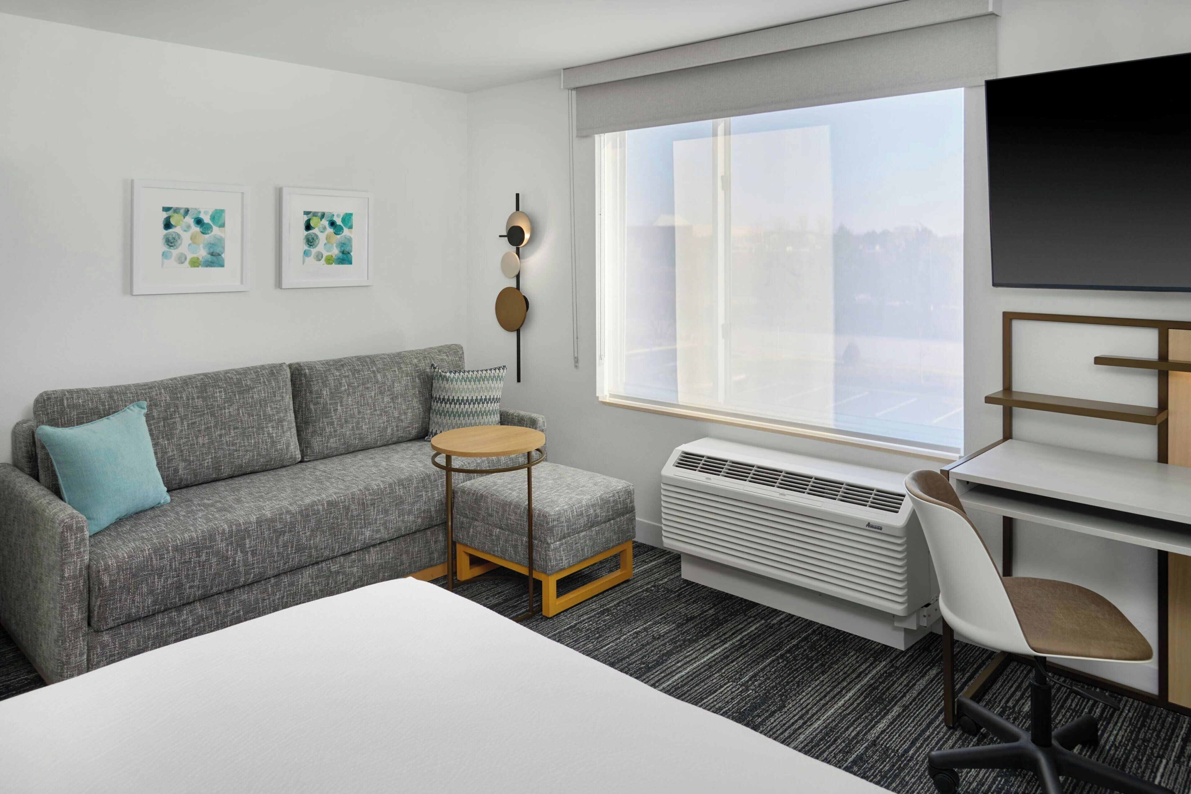 A modern hotel room with a grey sofa, a round table, a white desk, a chair, a TV, and a large window.
