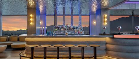 A bar area with a view of the sunset, a seating area with round tables, and a decorative ceiling.
