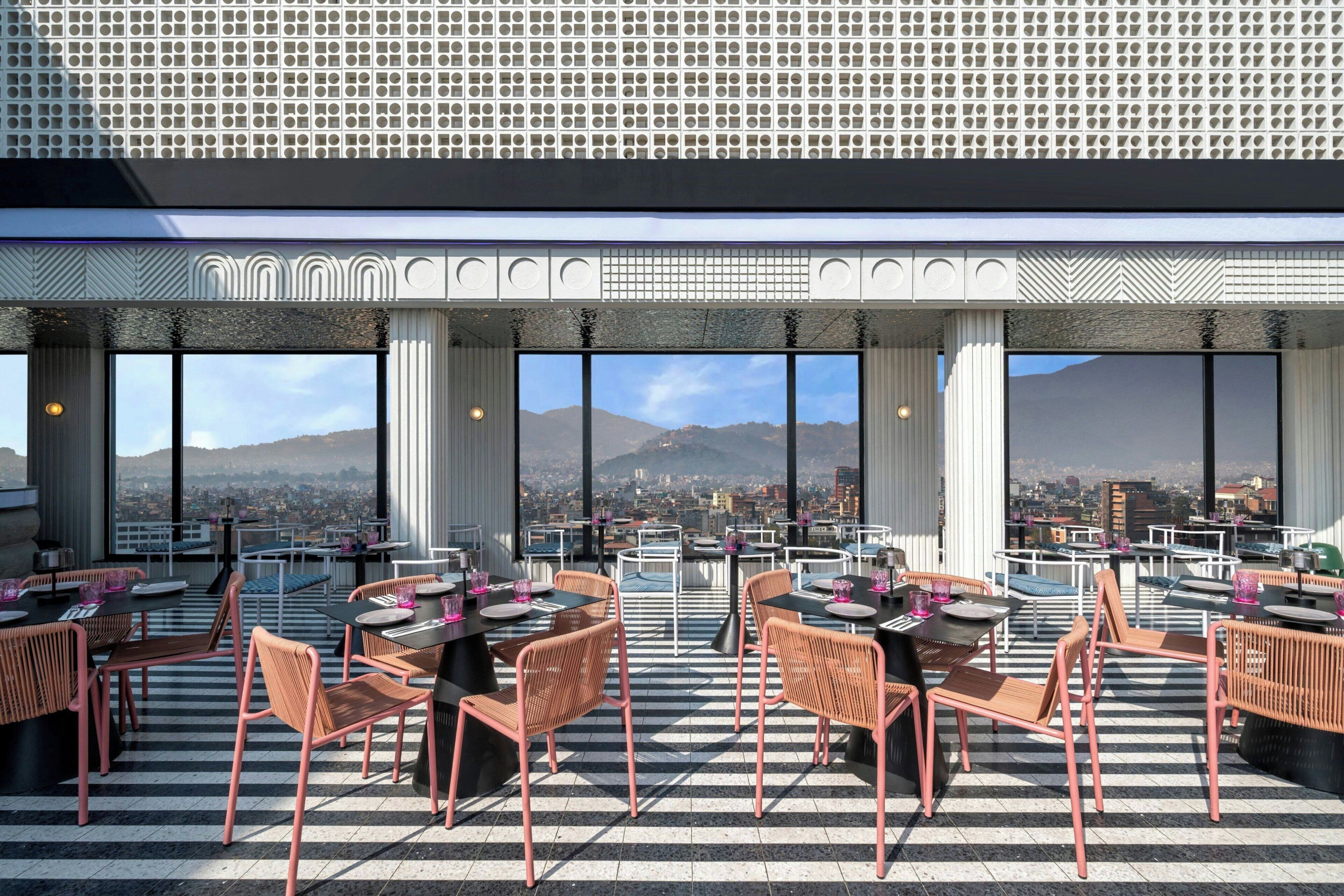 A rooftop dining area with tables and chairs, large windows, and a cityscape view.