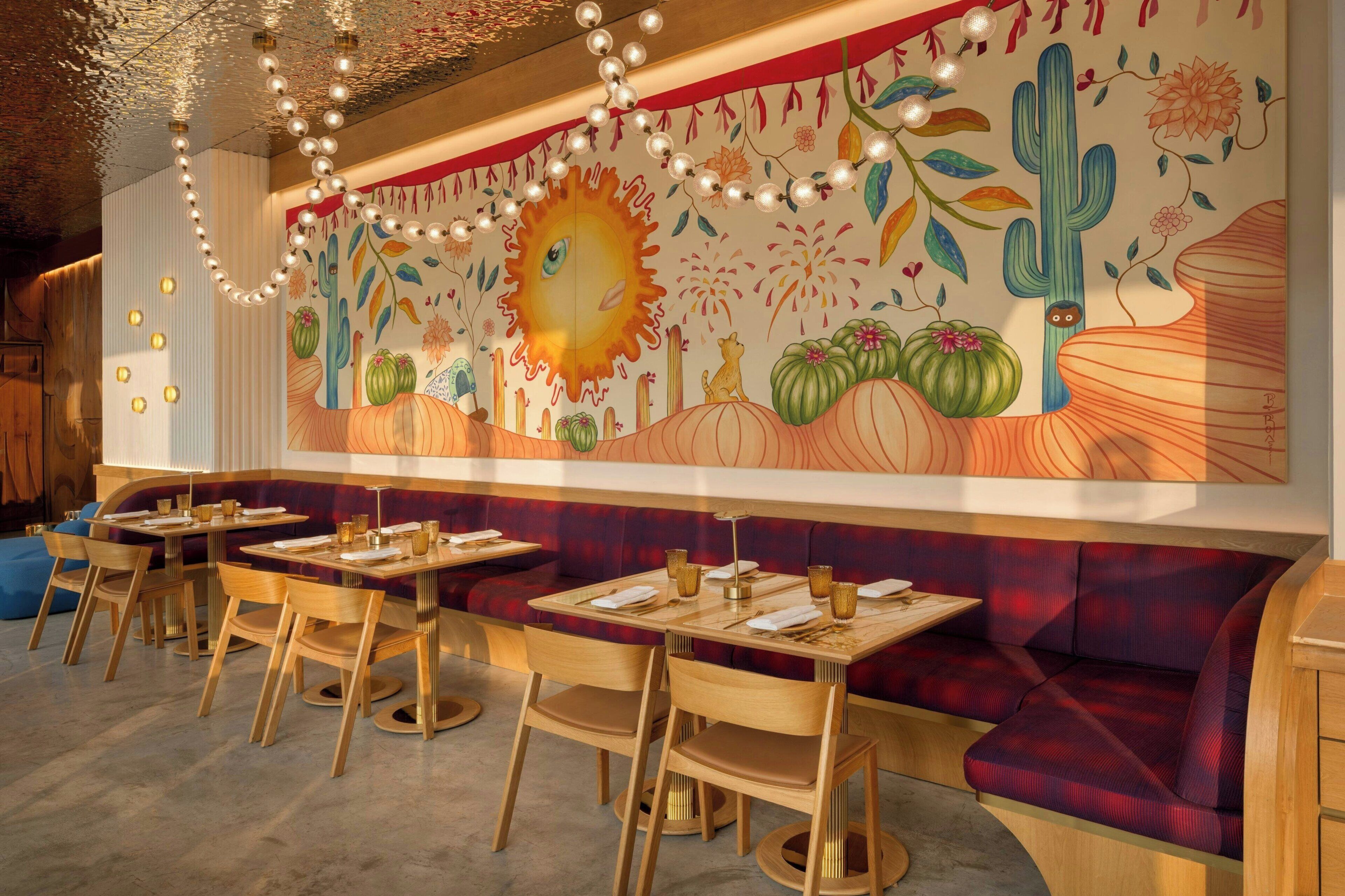 L’intérieur d’un restaurant est décoré de tables et de chaises en bois, d’une fresque représentant un soleil et des cactus, ainsi que d’un banc rouge rembourré.