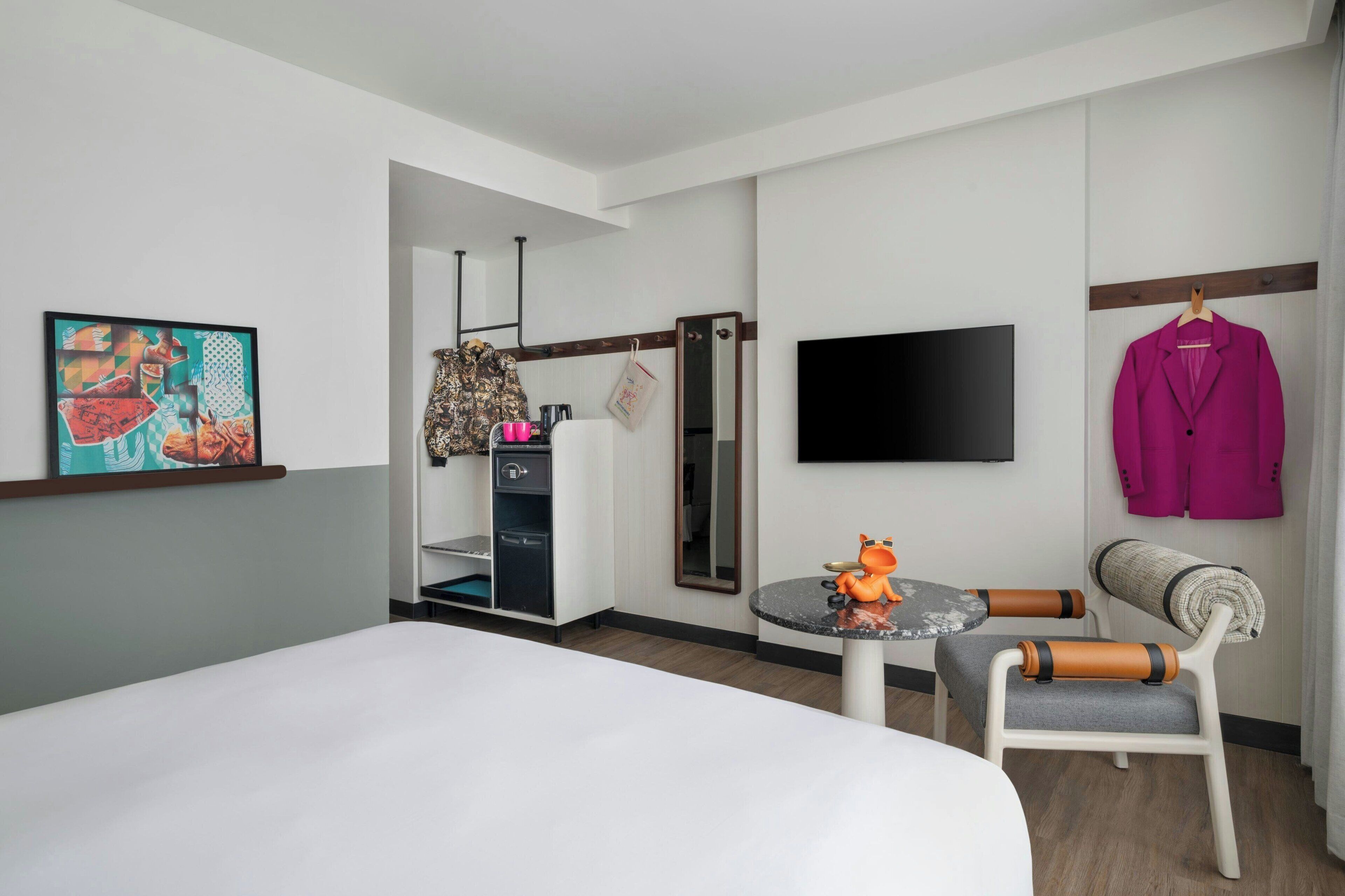 Ein Hotelzimmer mit einem Bett, einem kleinen Tisch mit einem Spielzeug, einem Stuhl, einem Fernseher und einem Kleiderhaken mit einem Mantel.