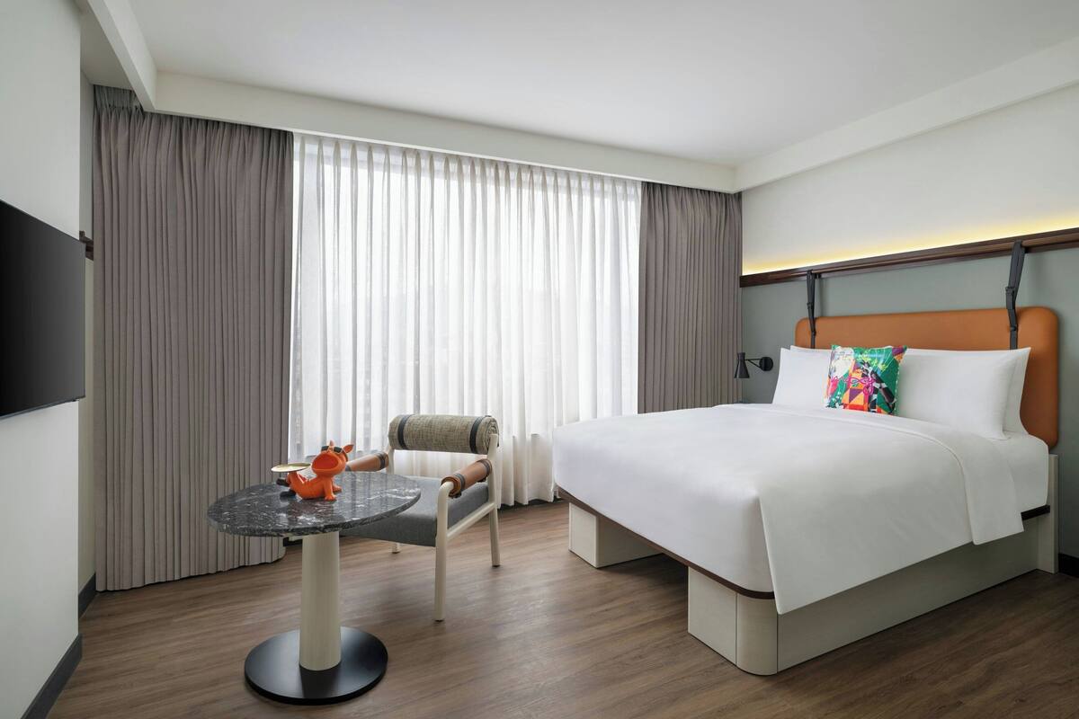 Habitación de hotel con cama, una mesa redonda con una silla, televisor montado en la pared y ventana con cortinas.
