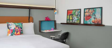 A hotel room with a bed, a chair, a shelf with books and a mug, and two framed artworks on the wall.