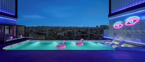 A rooftop pool with two pink flamingos, a cityscape view, and modern architecture with neon lights.