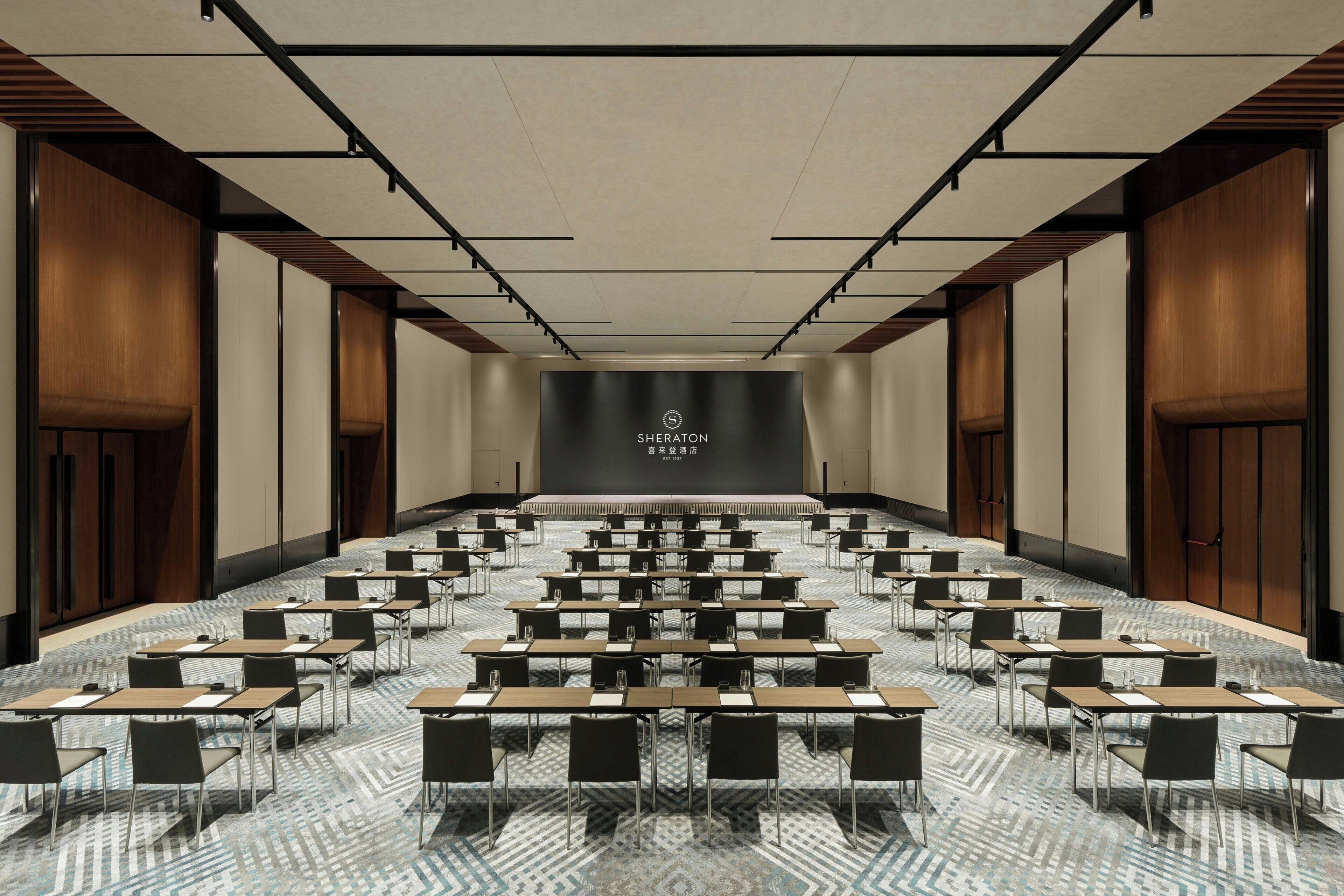 A conference room with rows of tables and chairs, a stage with a logo, and a patterned carpet.