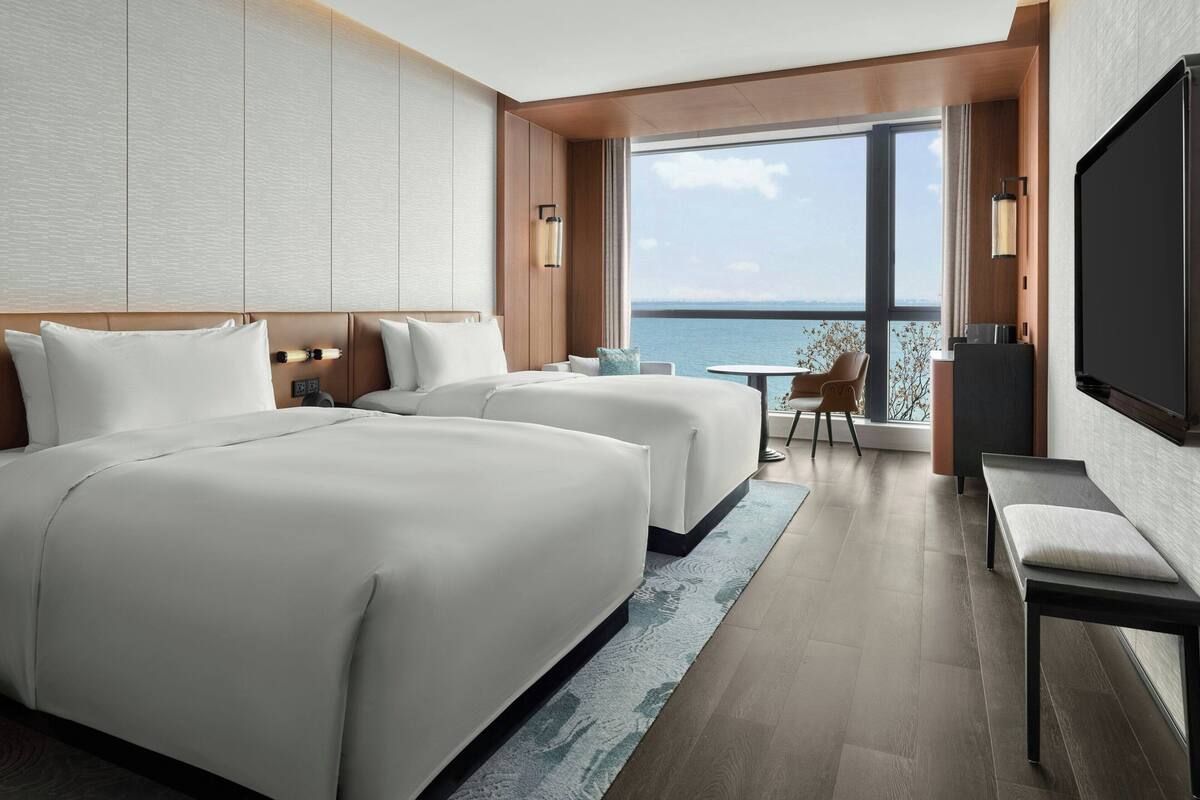 A hotel room with two beds, a TV, a balcony with a view of the sea, and a small table with chairs.