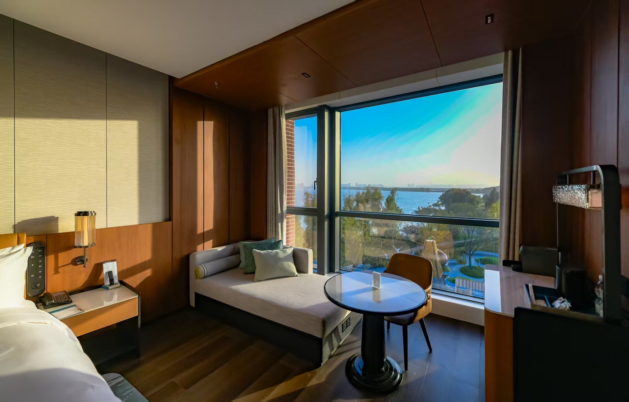 Habitación de hotel con cama, escritorio, silla, mesa redonda, ventana grande con vista al mar y televisión.