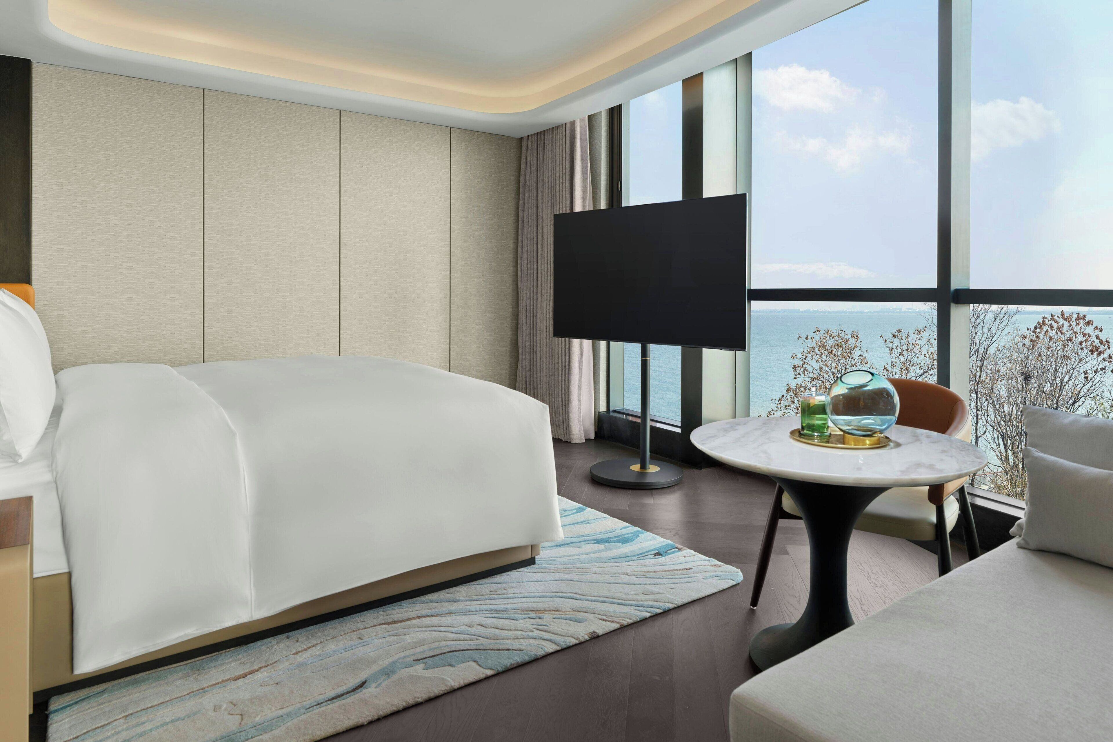 A modern hotel room with a large bed, a round table with a glass top, and a TV on a stand.