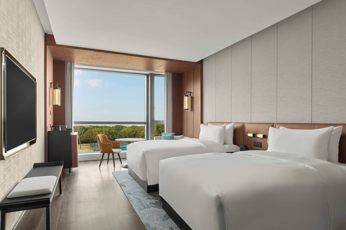 A hotel room with two beds, a TV, a desk, and a view of the outdoors.