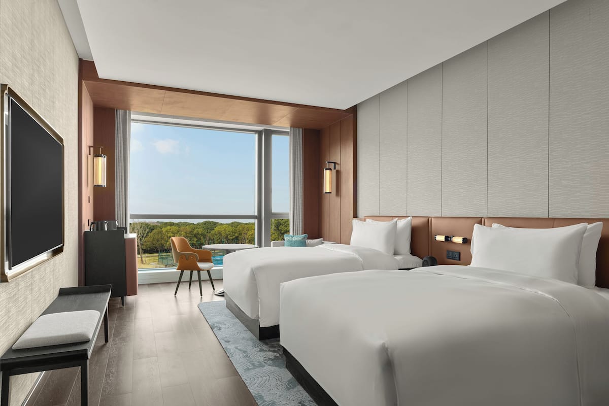 A hotel room with two beds, a TV, a desk, and a view of the outdoors.