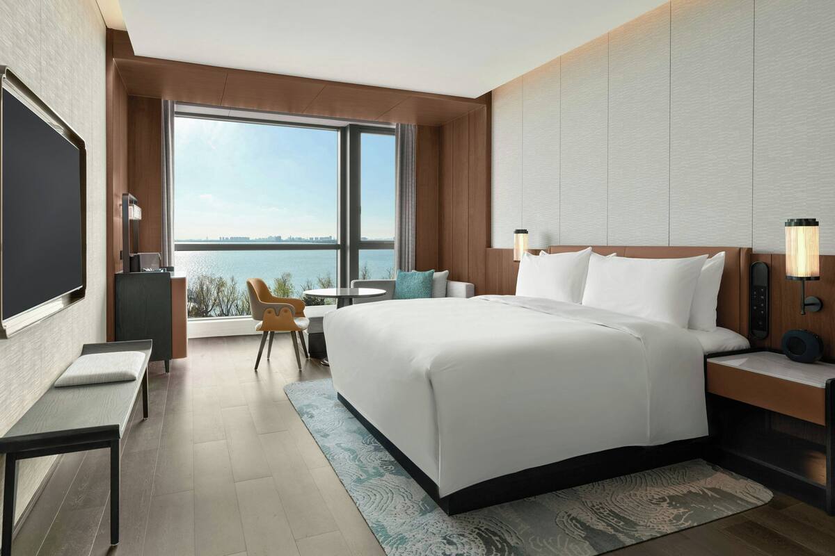 A modern hotel room with a large bed, a desk, a chair, a TV, and a view of the water.