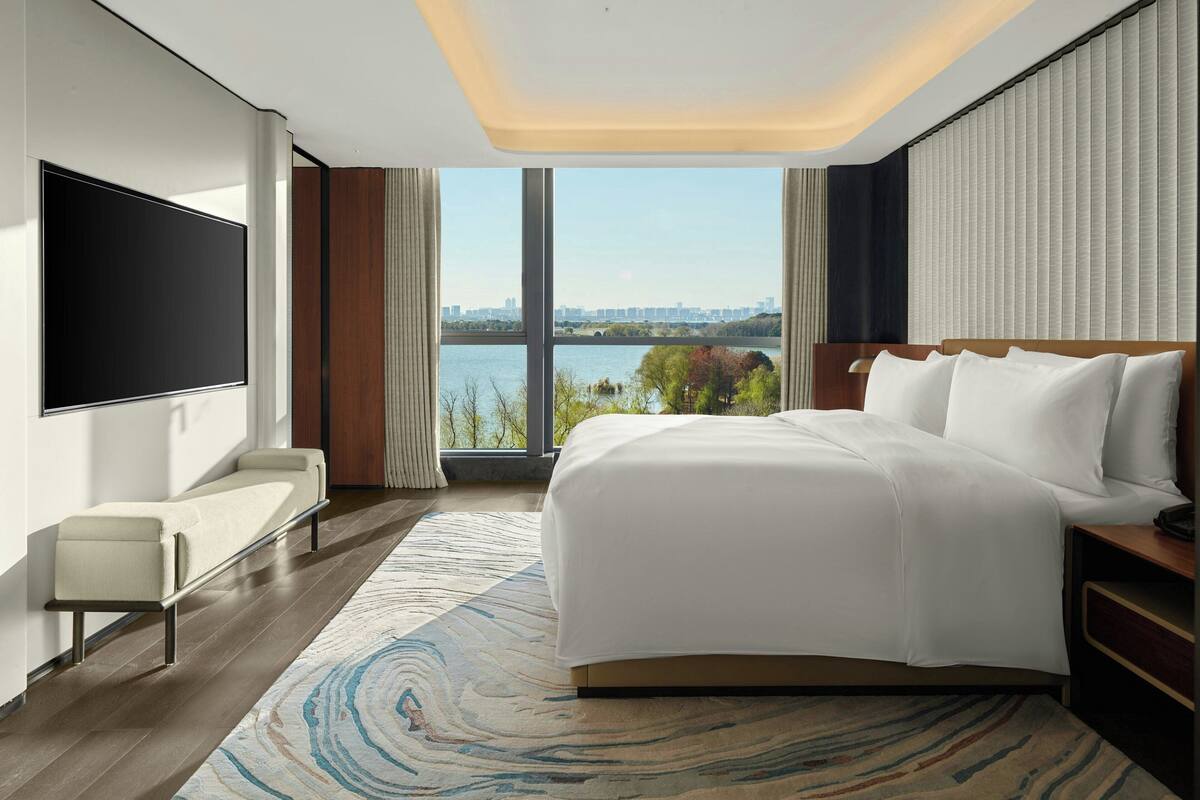 A modern hotel room with a large bed, a flat-screen TV, a seating area, and a view of a body of water.