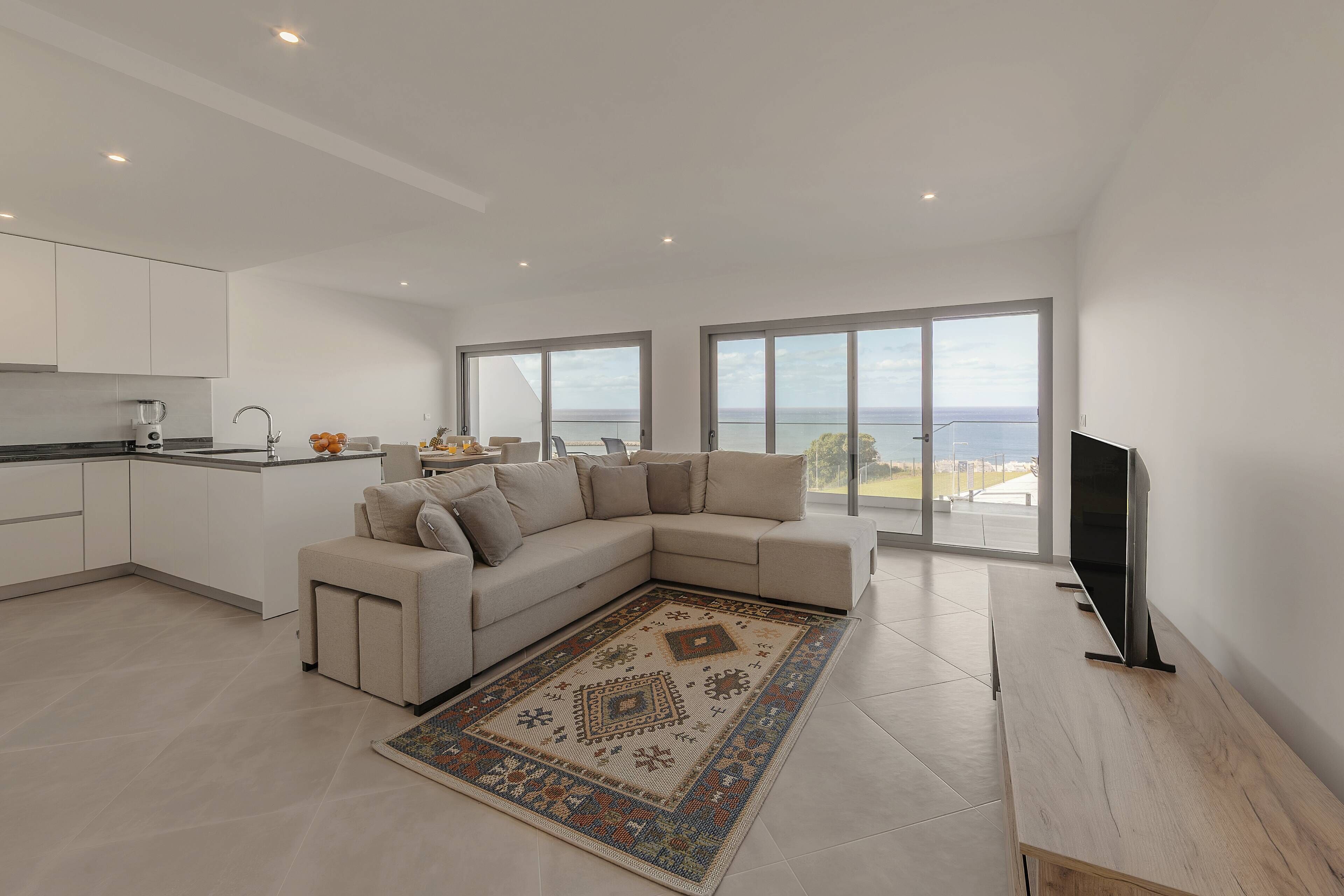 Un salon moderne avec un canapé d’angle beige, un téléviseur à écran plat et une grande fenêtre donnant sur la mer.