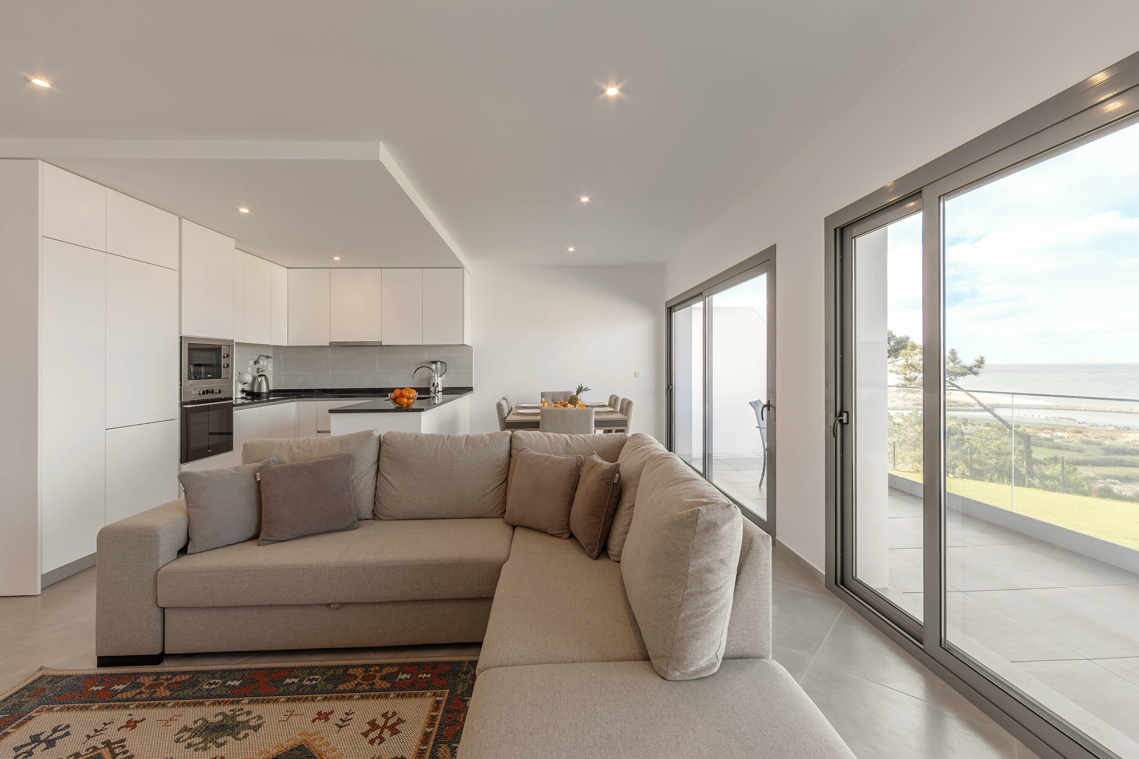 A modern living room with a beige sectional sofa, a kitchen area, and a large sliding glass door leading to a balcony with a view.