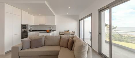 A modern living room with a beige sectional sofa, a kitchen area, and a large sliding glass door leading to a balcony with a view.
