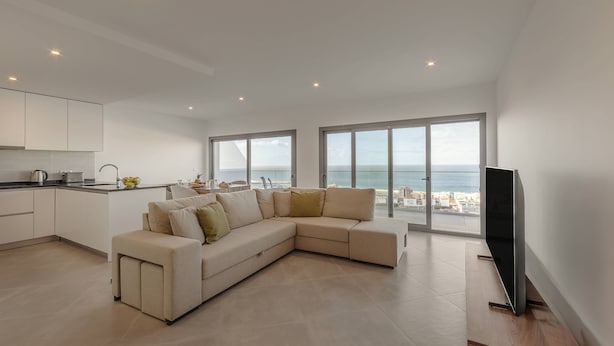 Un salon moderne doté d’un grand canapé en forme de L, d’un téléviseur à écran plat et offrant une vue sur la mer grâce à de grandes fenêtres.