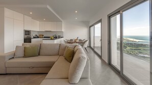 Un salon moderne avec un grand canapé d’angle, une cuisine en arrière-plan et un balcon offrant une vue sur la mer.