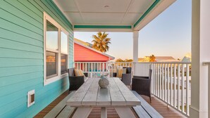 A covered porch with a wooden table and chairs, a white railing, and a view of a red building and palm trees in the background. - Good Gulf Times · New Listing! Heated Pool, Hot Tub & Steps to Beach (Gulf Shores)