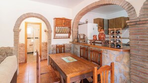 Un espace repas comprenant une table et des chaises en bois, un mur en pierre et un coin cuisine avec un réfrigérateur et un meuble à bouteilles.
