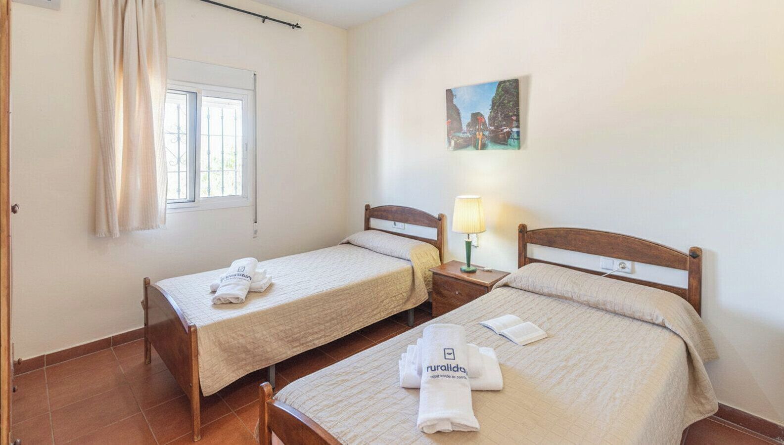 A room with two single beds, a window with curtains, a nightstand with a lamp, and a painting on the wall.