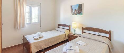 A room with two single beds, a window with curtains, a nightstand with a lamp, and a painting on the wall.