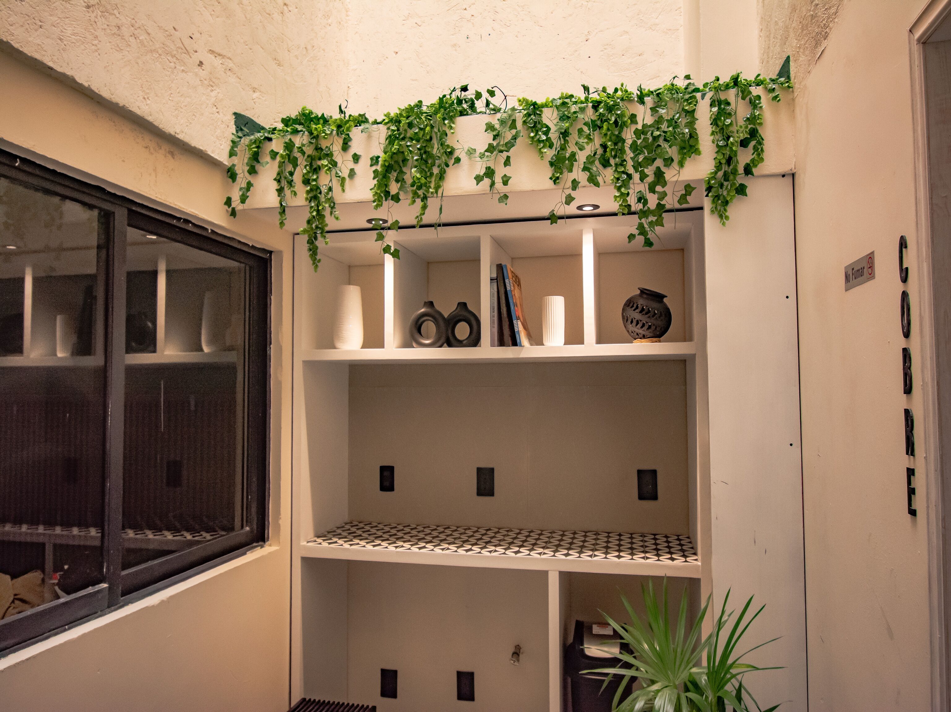 Een hoek van een kamer met een witte muur, een raam met een zwarte omlijsting en een wandplank met decoratieve objecten en een plant.