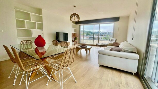 Een moderne woonkamer met een glazen eettafel, rieten stoelen, een witte bank en een groot raam met uitzicht op de stad.